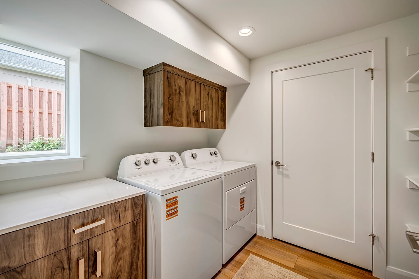 Side-by-side washer and dryer with cabinet above, counter adjacent with a window above, and a door on the wall.