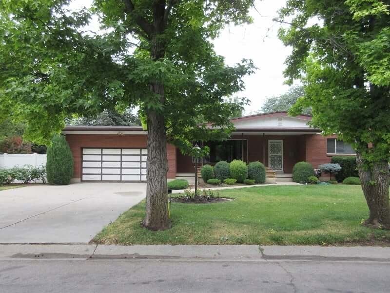 Ranch Rambler style house before the exterior makeover. The landscaping includes two trees in the front, green grass, and various hedges closer to the house. The red brick house includes a two-car garage with a wide driveway in front and a covered front porch.
