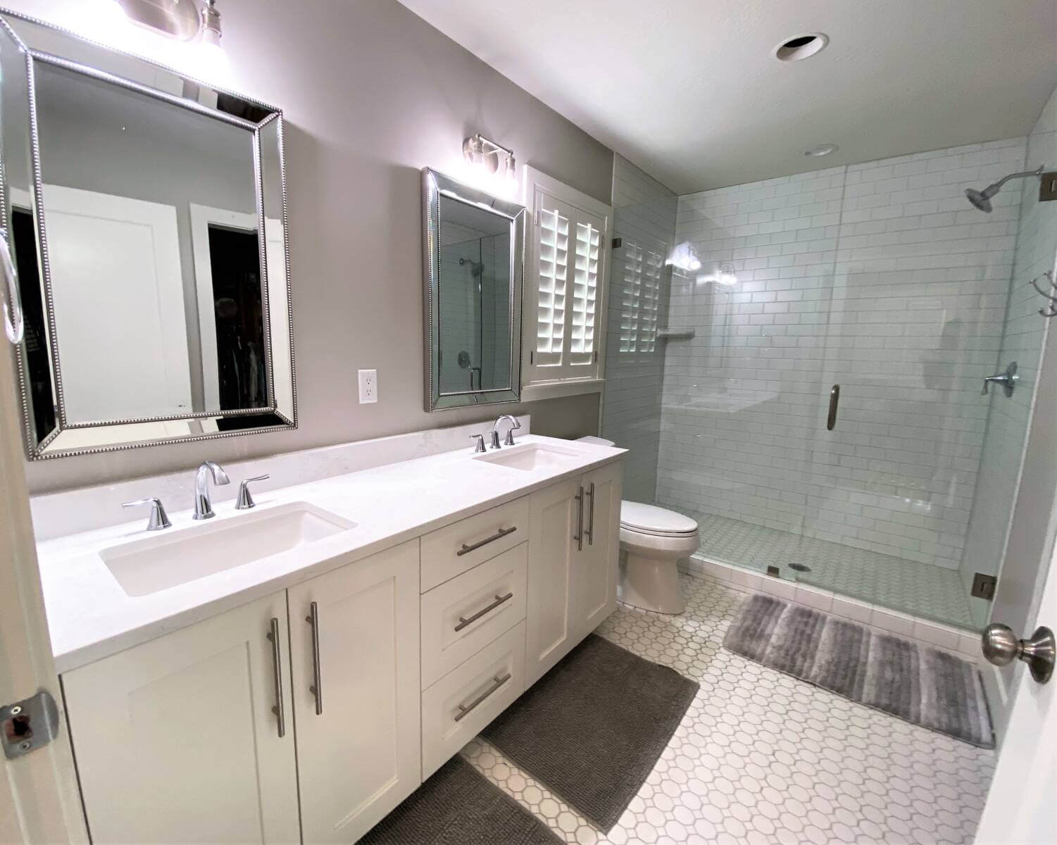 Primary bathroom remodel with two sinks, a toilet under the window, and a large shower. On the floor are three bath mats.