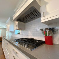 Thumbnail of http://Kitchen%20stove%20with%20white%20cabinetry%20and%20a%20hood%20above.%20Next%20to%20the%20stove%20is%20a%20large%20red%20jar%20with%20cooking%20utensils.