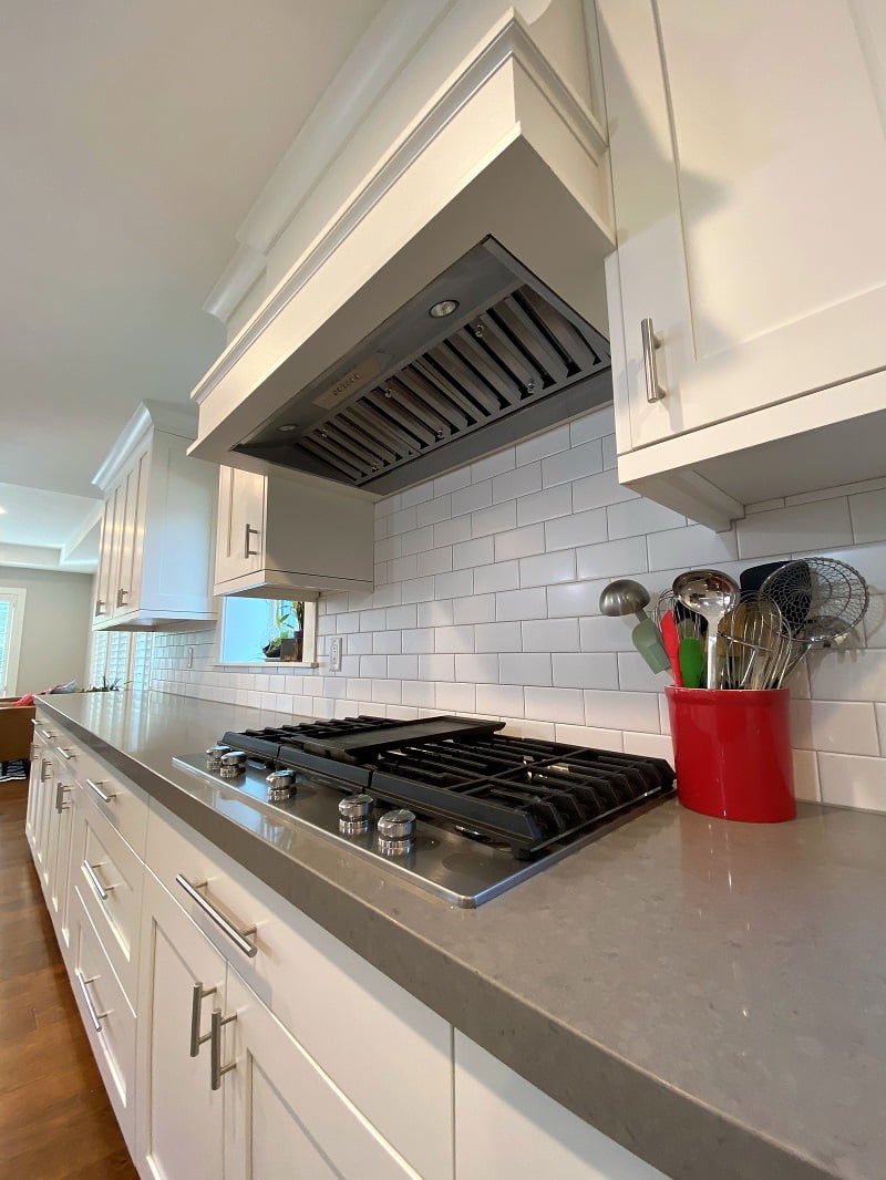 Kitchen stove with white cabinetry and a hood above. Next to the stove is a large red jar with cooking utensils.