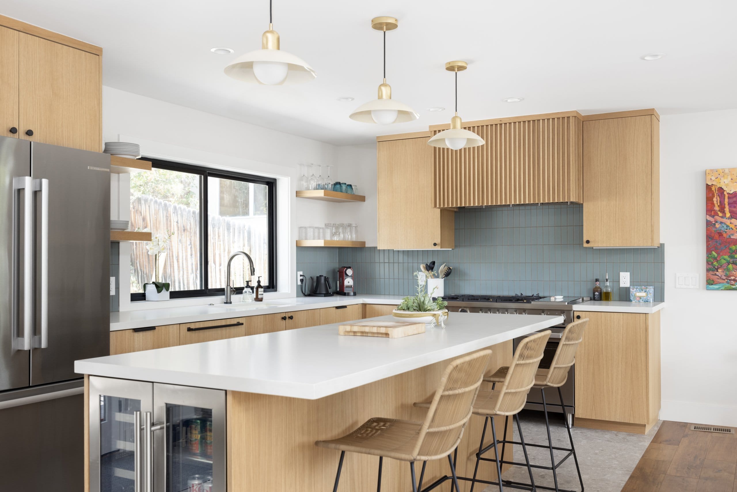 Updated modern and colorful kitchen with the sink placed under the large window, wood cabinetry, terrazzo tile floors, and an island in the center.
