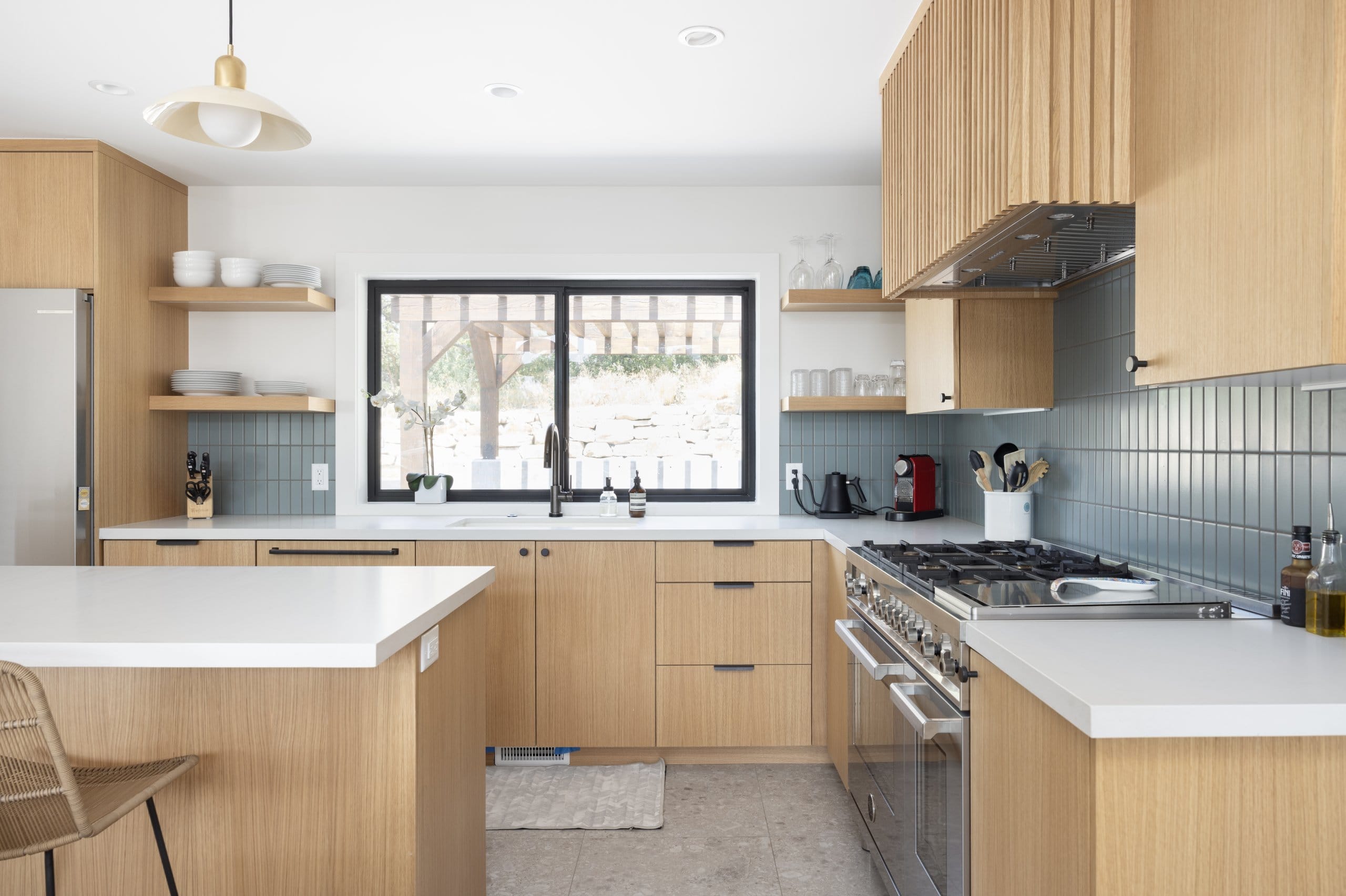 Updated modern and colorful kitchen with the sink placed under the large window, wood cabinetry, terrazzo tile floors, and an island in the center.