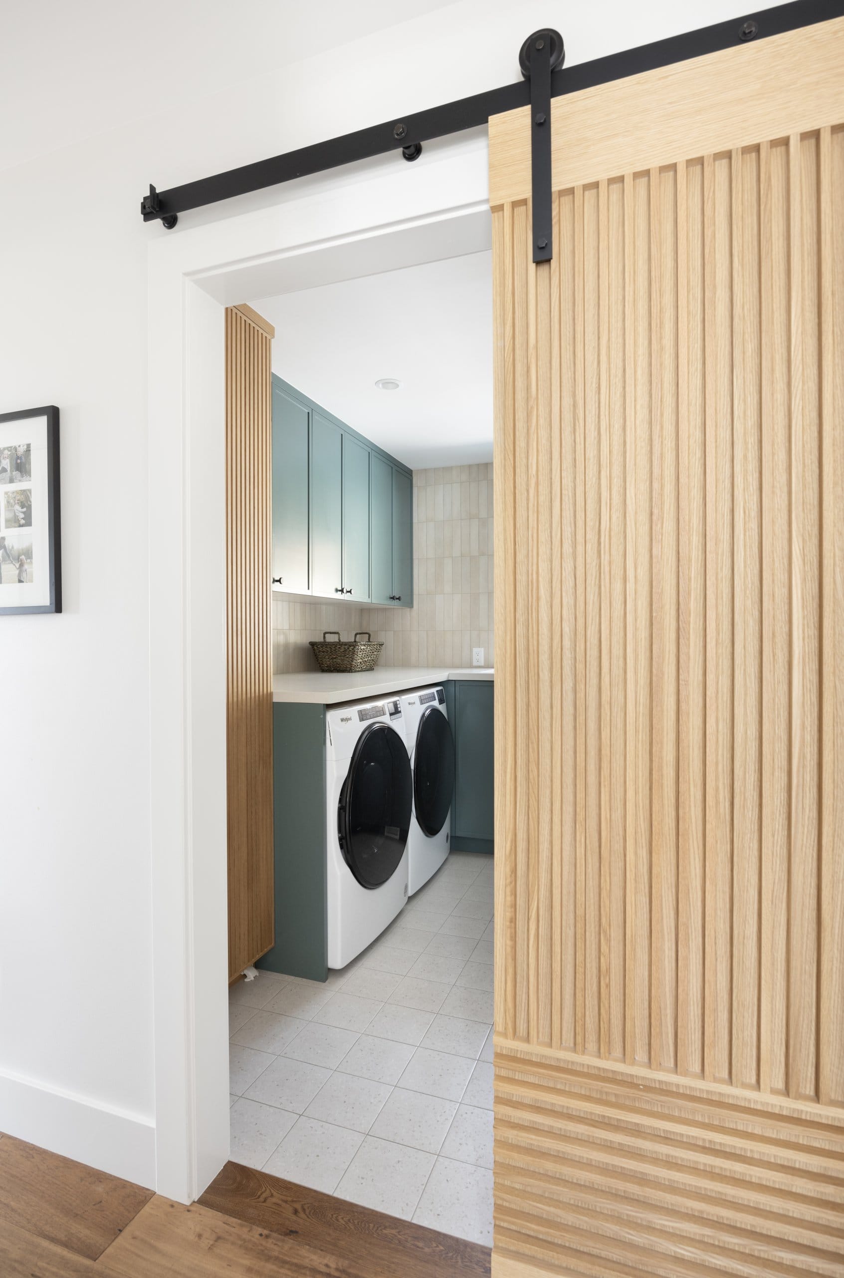 Wood barn door part way open with view into the large laundry room. The laundry room features cabinetry painted in a fun color, a folding counter above the side-by-side washer and dryer, and a spacious layout.