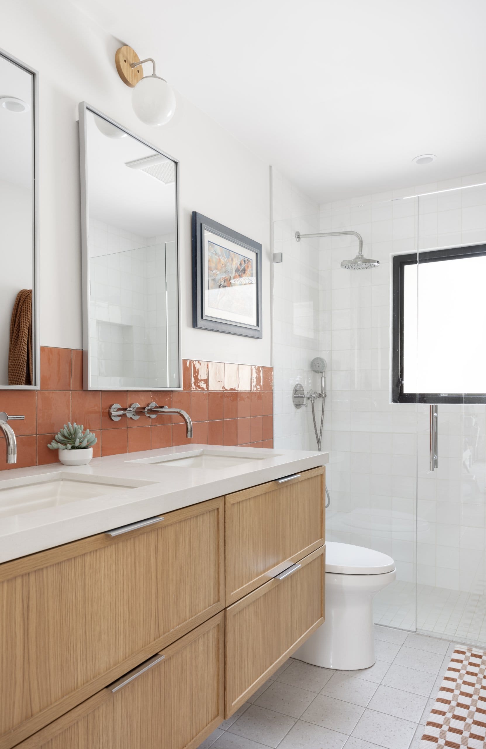 Modern bathroom with double sinks, toilet, and a window in the shower. The tile is neutral on the floor and in the shower with a fun pop of orange tile behind the vanity.