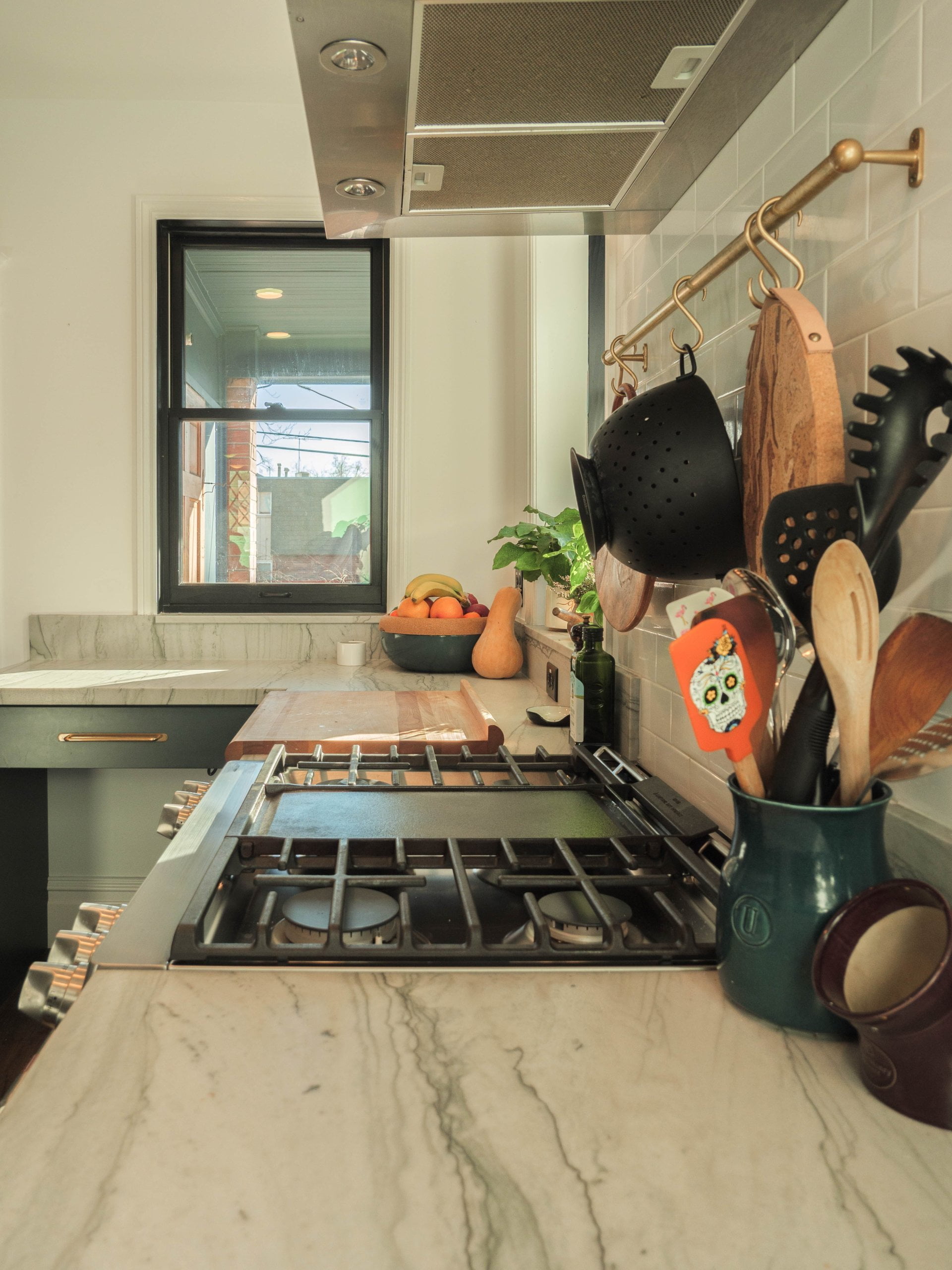 Historic home kitchen remodel looking across the stove to the window. On the counter is a jar with cooking utensils, a cutting board, and a bowl of fruit.