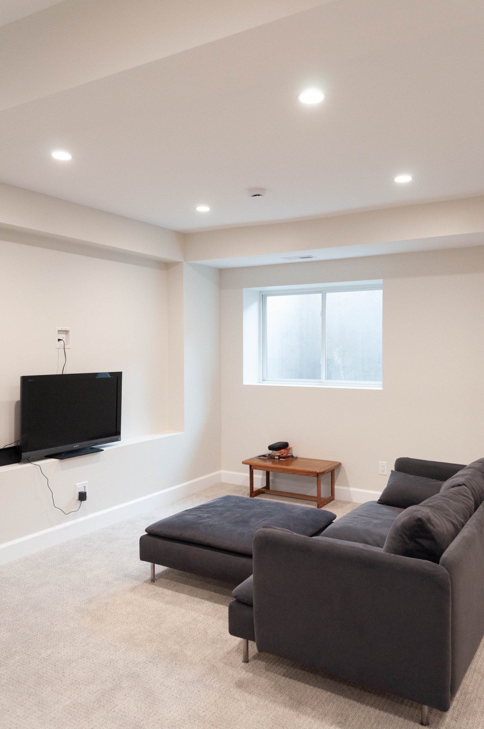 Basement family room after the remodel with a couch and ottoman, bench under the egress window, and a TV.
