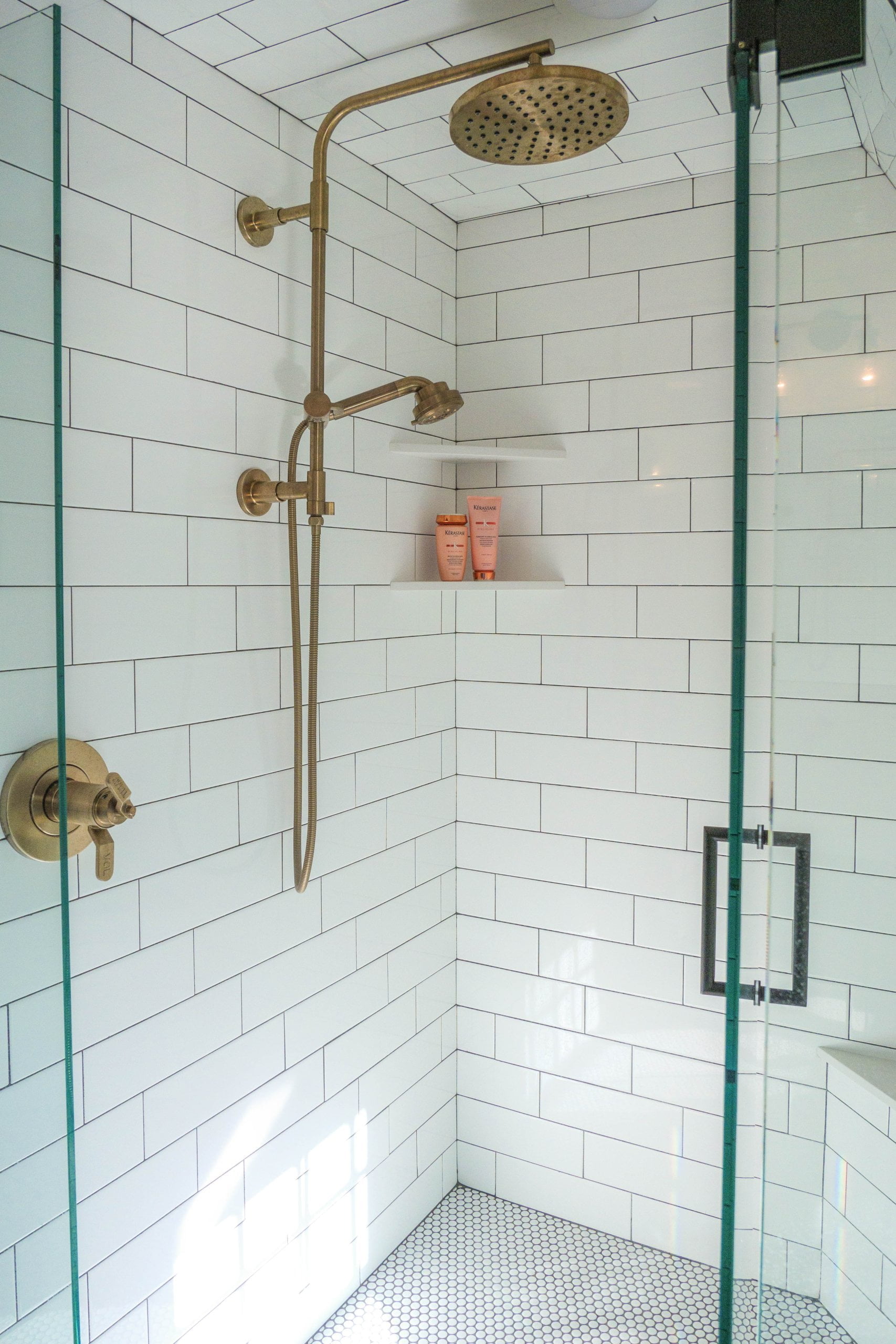 Shower with glass door, shelves for soaps, and a showerhead with a detachable part. The tile in the shower is white with subway tile on the ceiling and walls.
