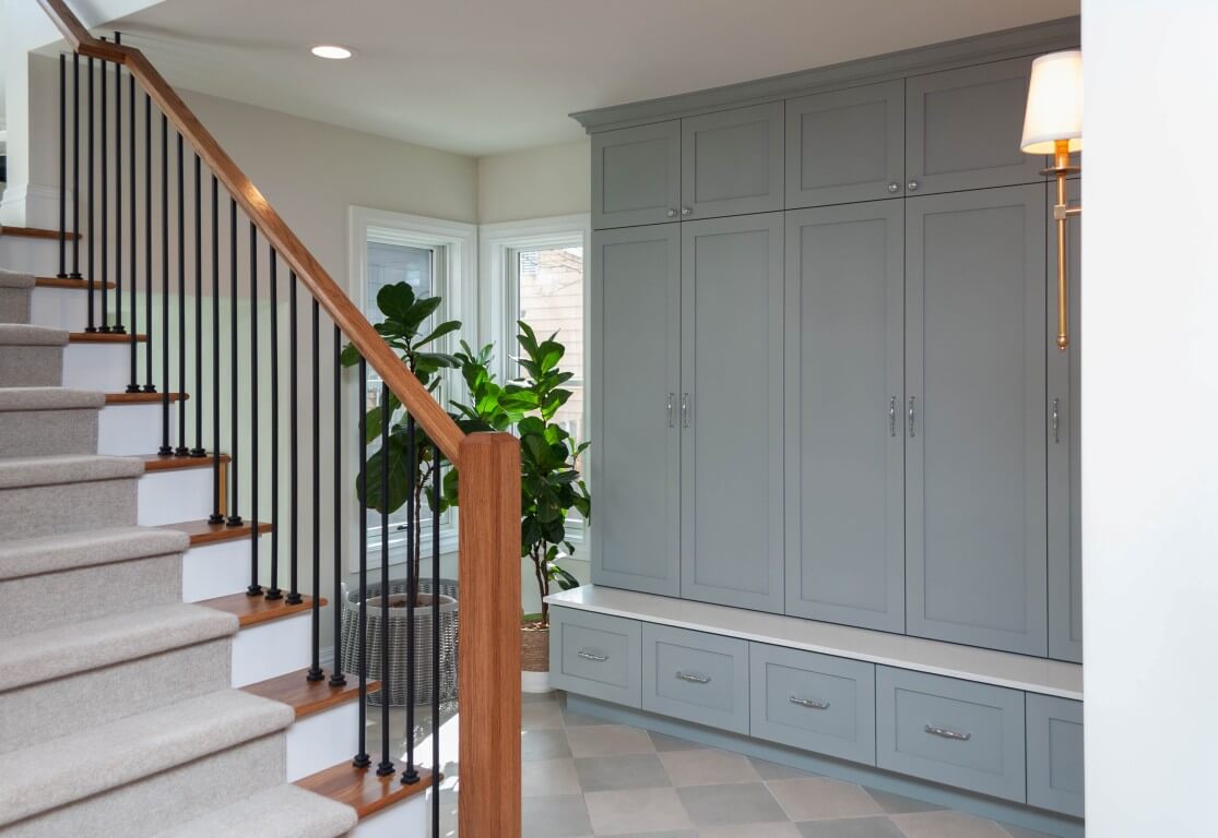 A staircase with a mudroom on the other side of the railing. The mudroom includes large built-in cabinets, plants, and tile flooring.