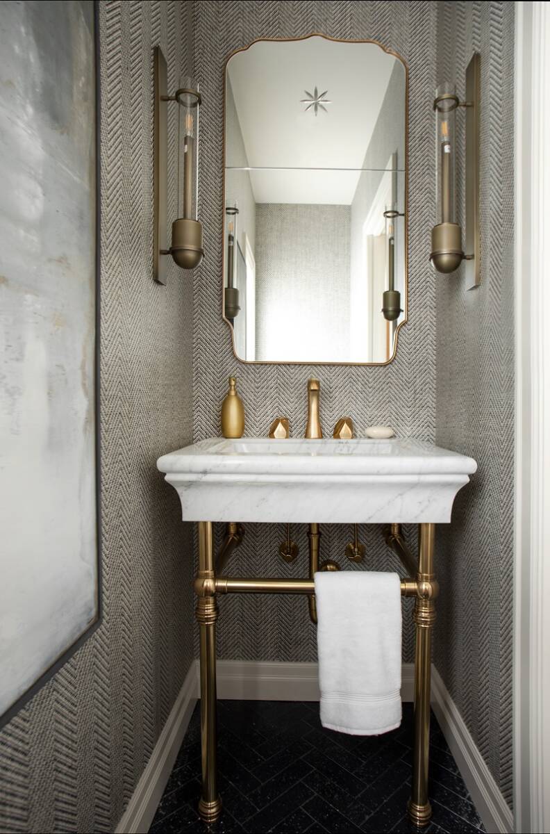 Vanity in the powder bathroom with gold fixtures, wallpaper, and a hand towel hung under the open sink.
