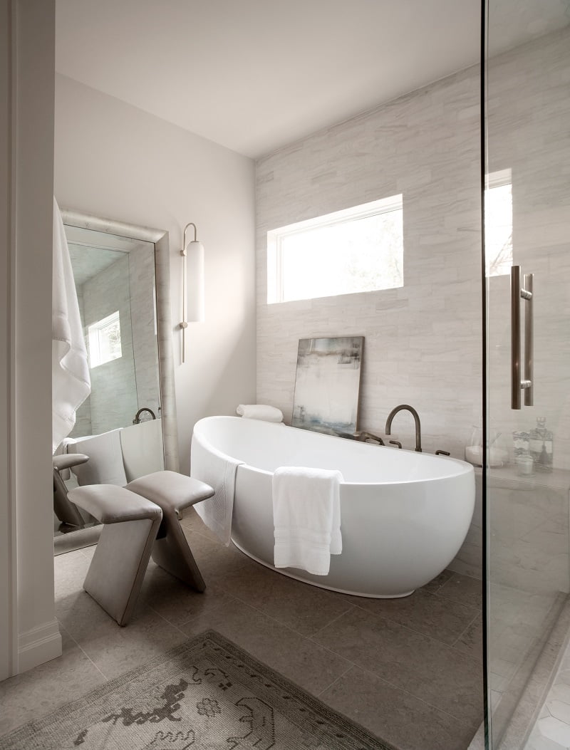 Large freestanding tub in the primary bathroom. Along the wall is a bench with various decorations including a painting. On the other wall is a large mirror with a stool in front.