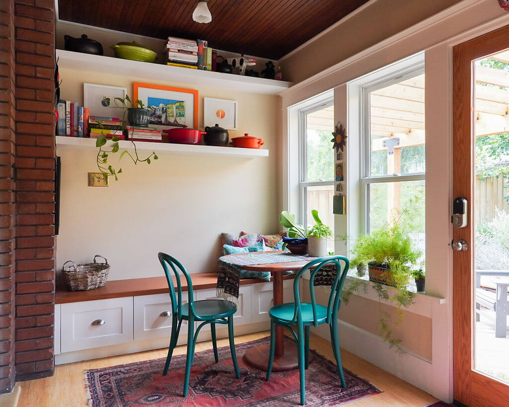 A sitting area in a small addition with a built-in bench, round table, and two chairs. The space also includes shelves on the wall and lots of natural light from the windows.
