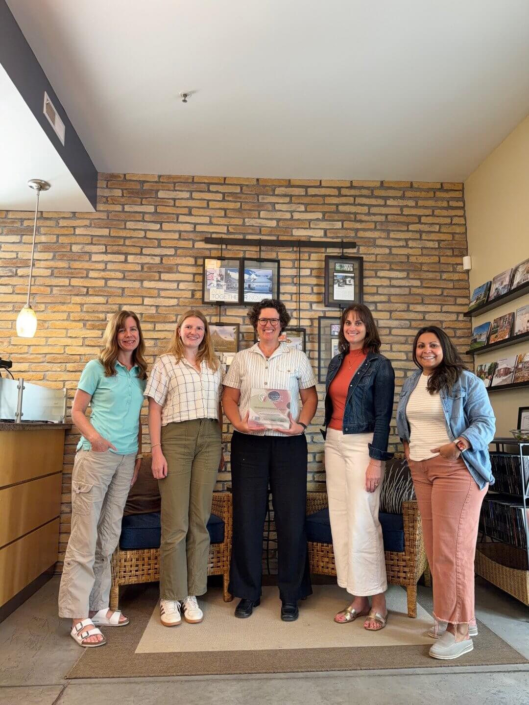 Group photo with five people standing in front of a brick wall. The person in the center is holding an award. Recognition.