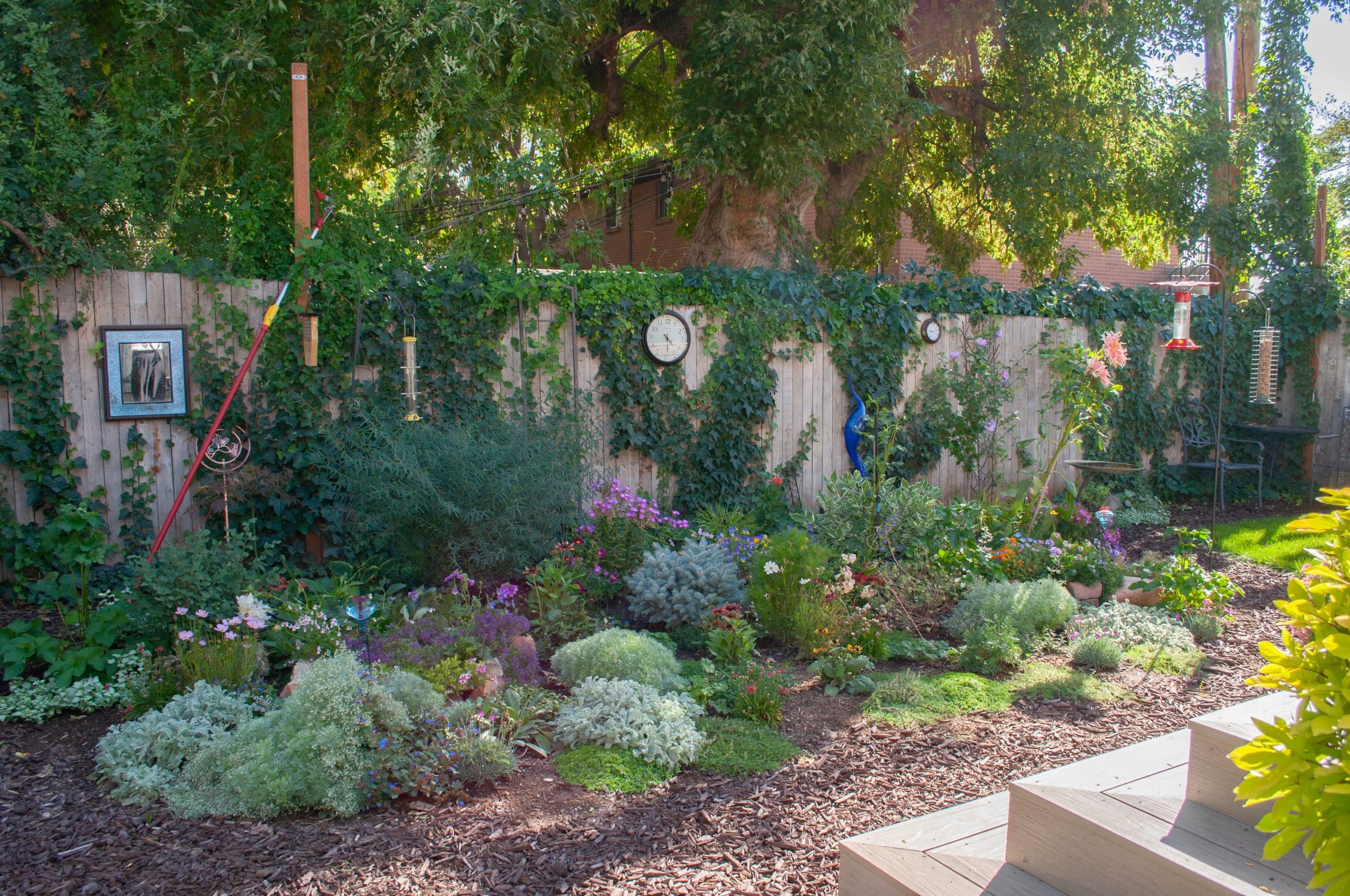 Small backyard landscaping with flowers planted in mulch, a wood fence a hummingbird feeder, and a small view of the new steps leading to the back door.