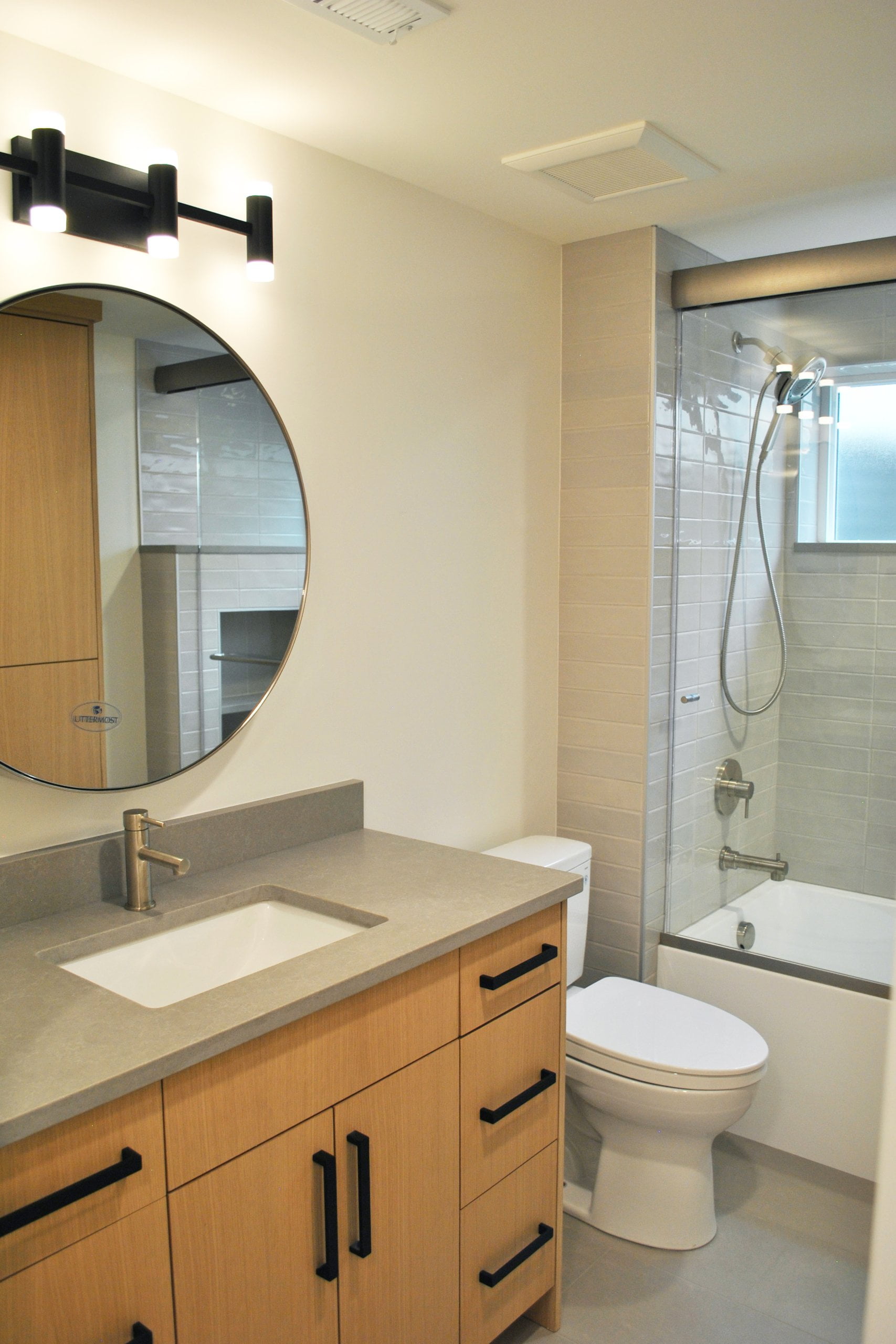 Bathroom renovation with a sink, toilet, and combined tub and shower. The sink has cabinets and drawers underneath and a round mirror and light fixture hung above.