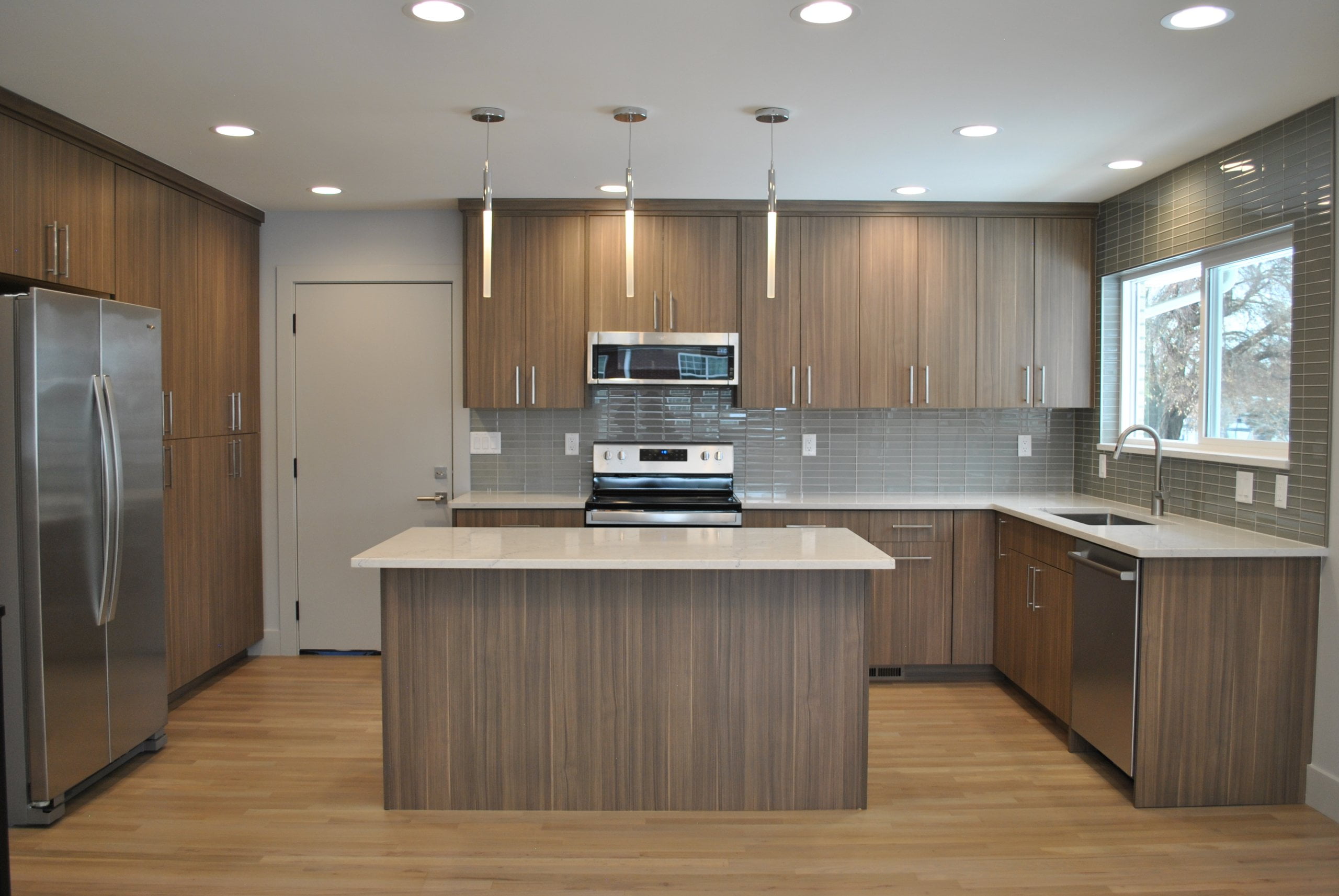 Large kitchen with the sink centered under the window on the wall to the right, stove and oven straight ahead, and the fridge and pantry cabinets to the left. In the center is an island with room for stools.