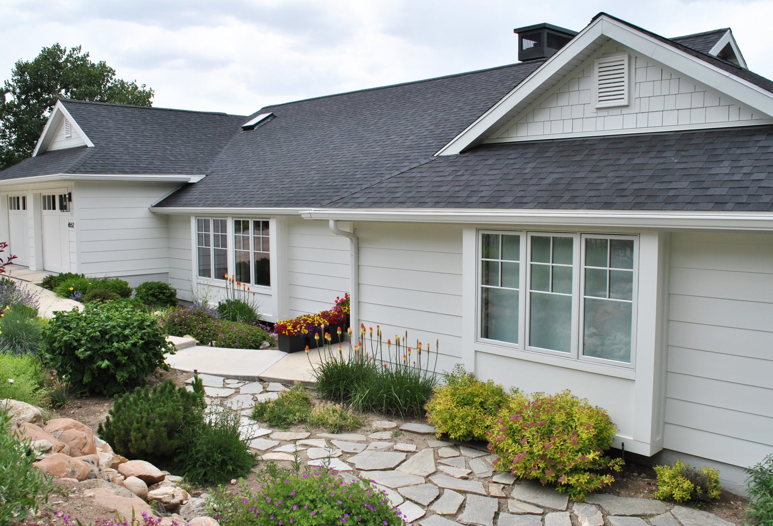 Exterior of house after the remodel. The house includes two garage doors, flowers in the landscaping, white siding and windows, and a small stone walkway.