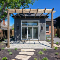 Thumbnail of http://Exterior%20of%20the%20addition%20with%20a%20pergola%20and%20patio,%20large%20windows,%20and%20gray%20cement%20siding.