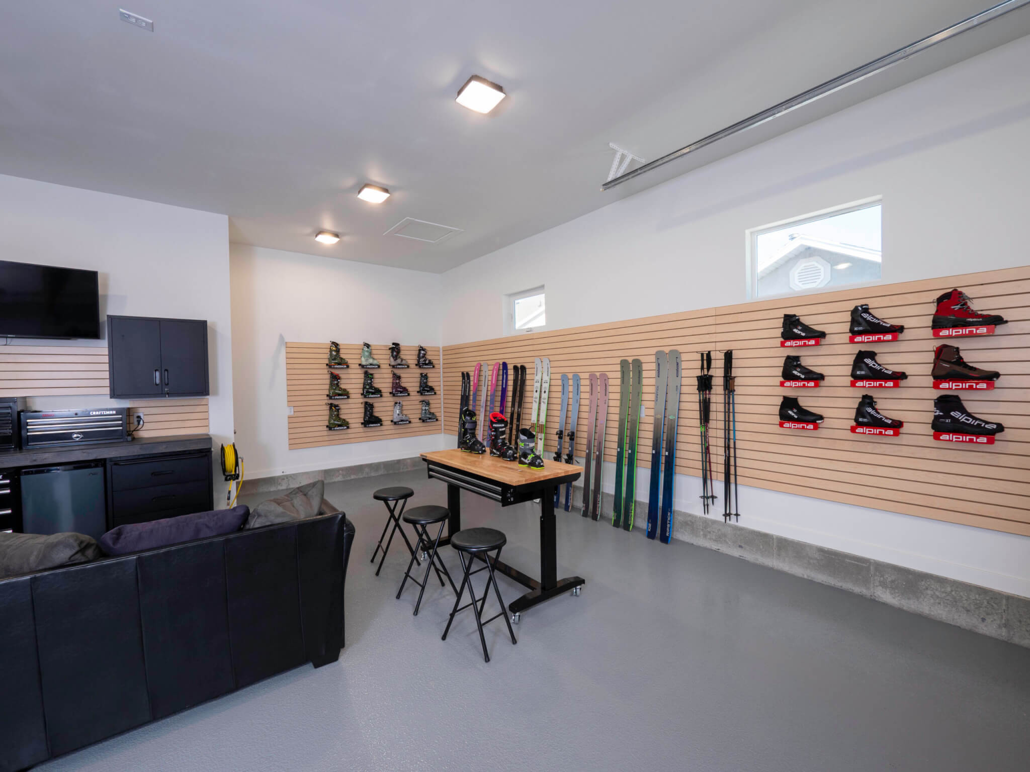 Garage slatwall with storage for skis, boots, and poles. The wall includes two windows and a rolling table is placed in the center with stools and more ski boots on top.