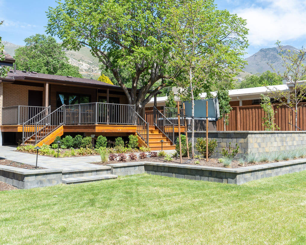 Backyard of a mid-century home. The yard features grass, a patio with a firepit, a hot tub, and a deck built around a tree.