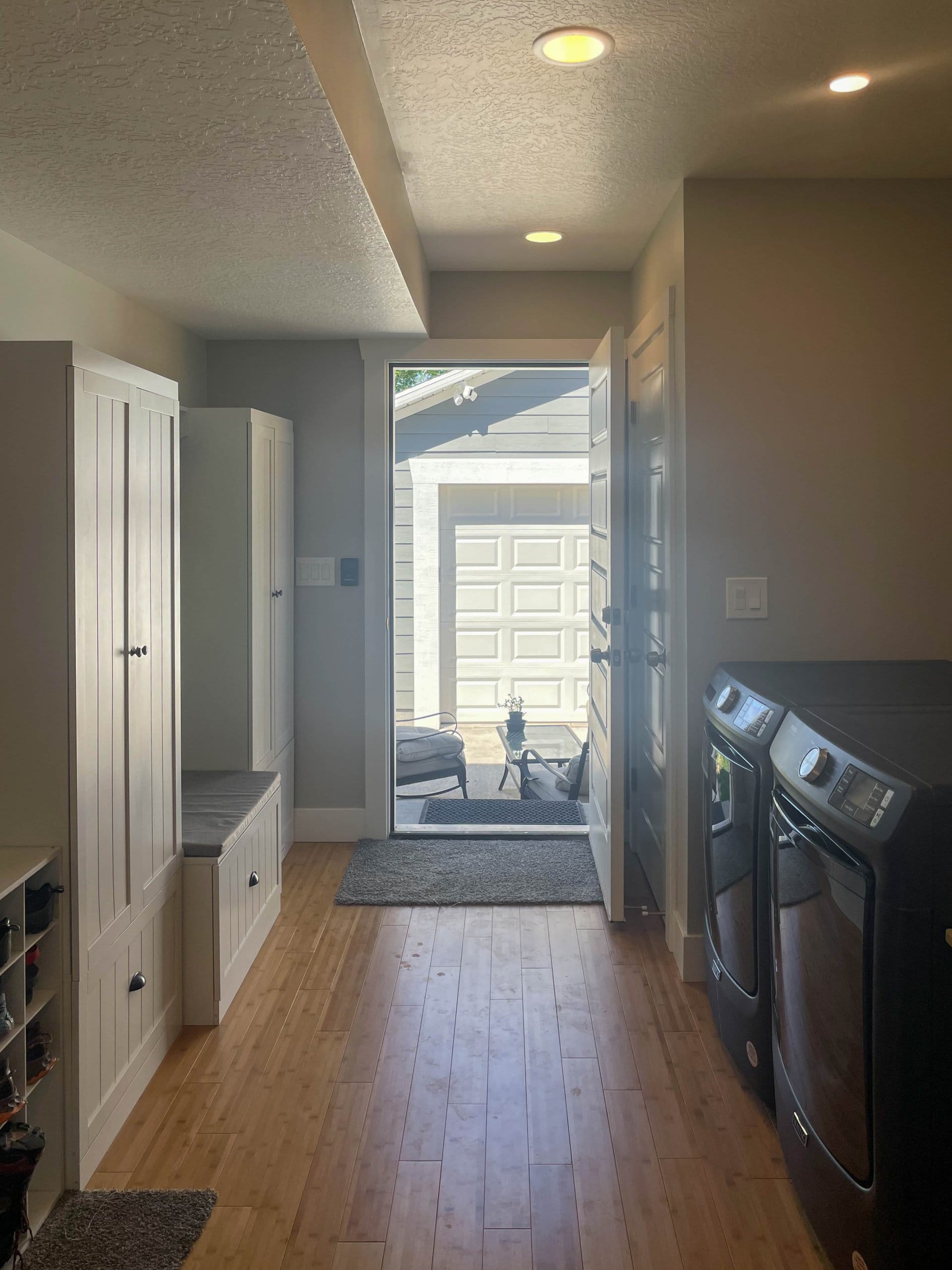 Combined mudroom and laundry room with washer and dryer placed side-by-side to the right and cabinets to the left. Door to the backyard is open showing the patio furniture in front of the garage.
