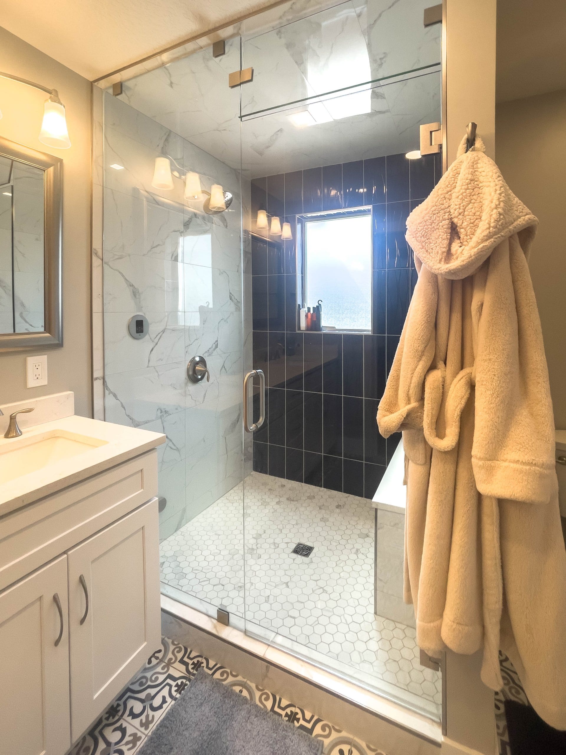 Large shower with a window and bench. Outside the glass door is a robe hung on the wall and a partial view of the sink.