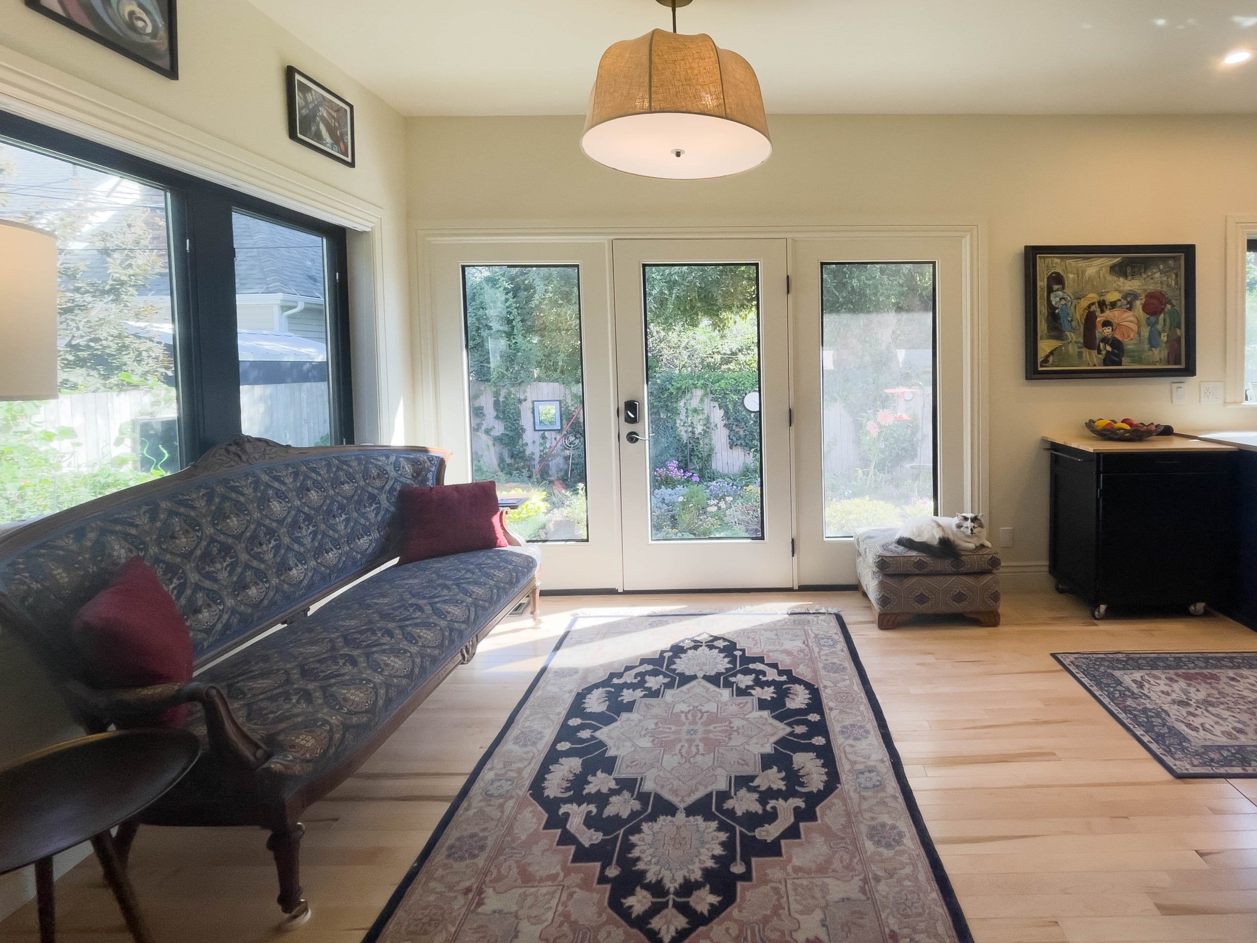 Door to the backyard with windows on either side and on the wall to the left. The rooms furniture incudes a rug in the center, a couch along the wall to the right, pictures hung on the walls, and an ottoman in front of one of the windows that a cat is sitting on.