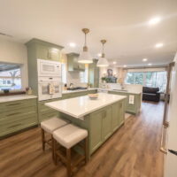 Thumbnail of http://Large%20kitchen%20with%20green%20cabinets,%20gold%20hardware,%20and%20white%20appliances.%20In%20the%20middle%20is%20a%20large%20island%20with%20seating%20and%20lights%20hung%20above.