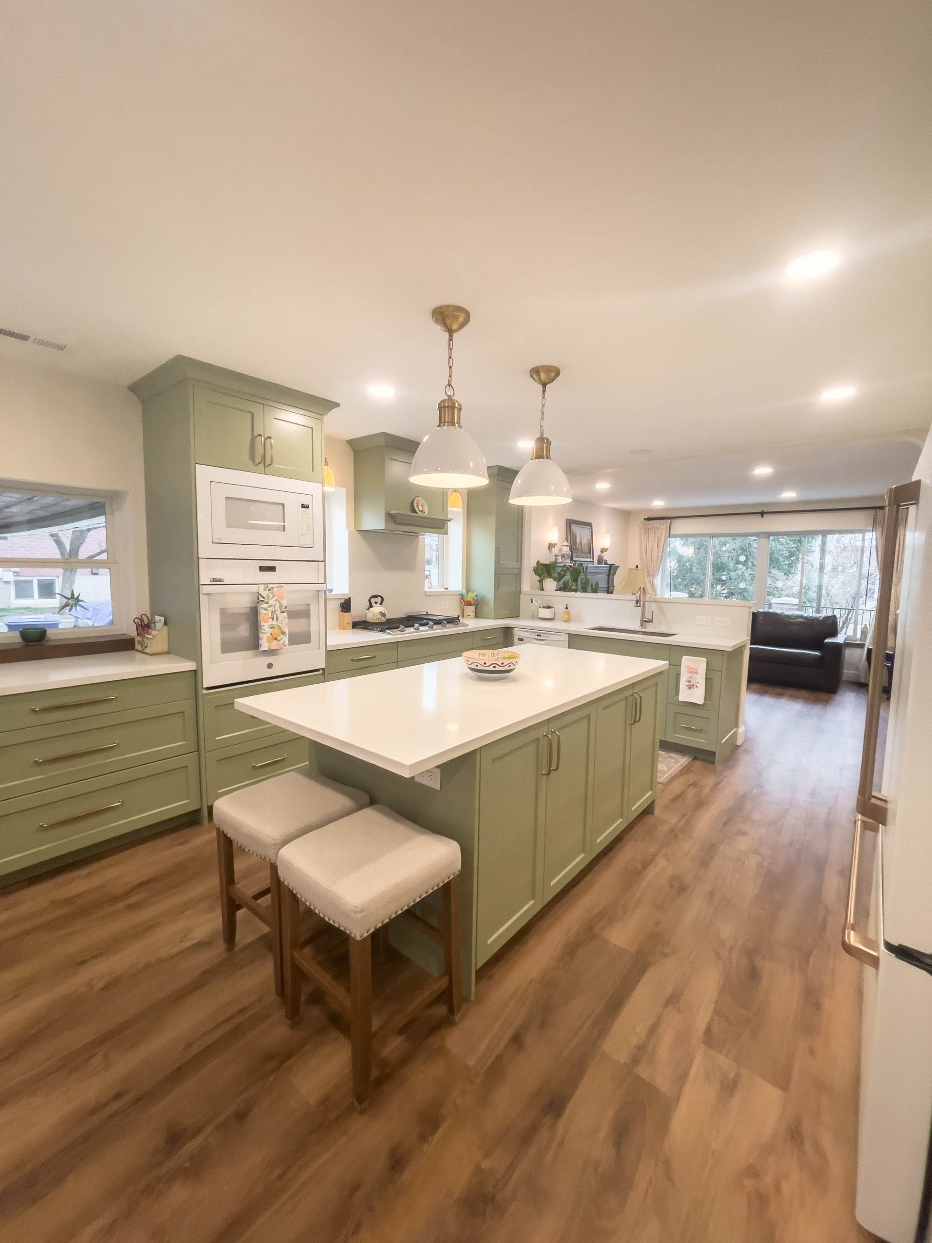 Large kitchen with green cabinets, gold hardware, and white appliances. In the middle is a large island with seating and lights hung above.