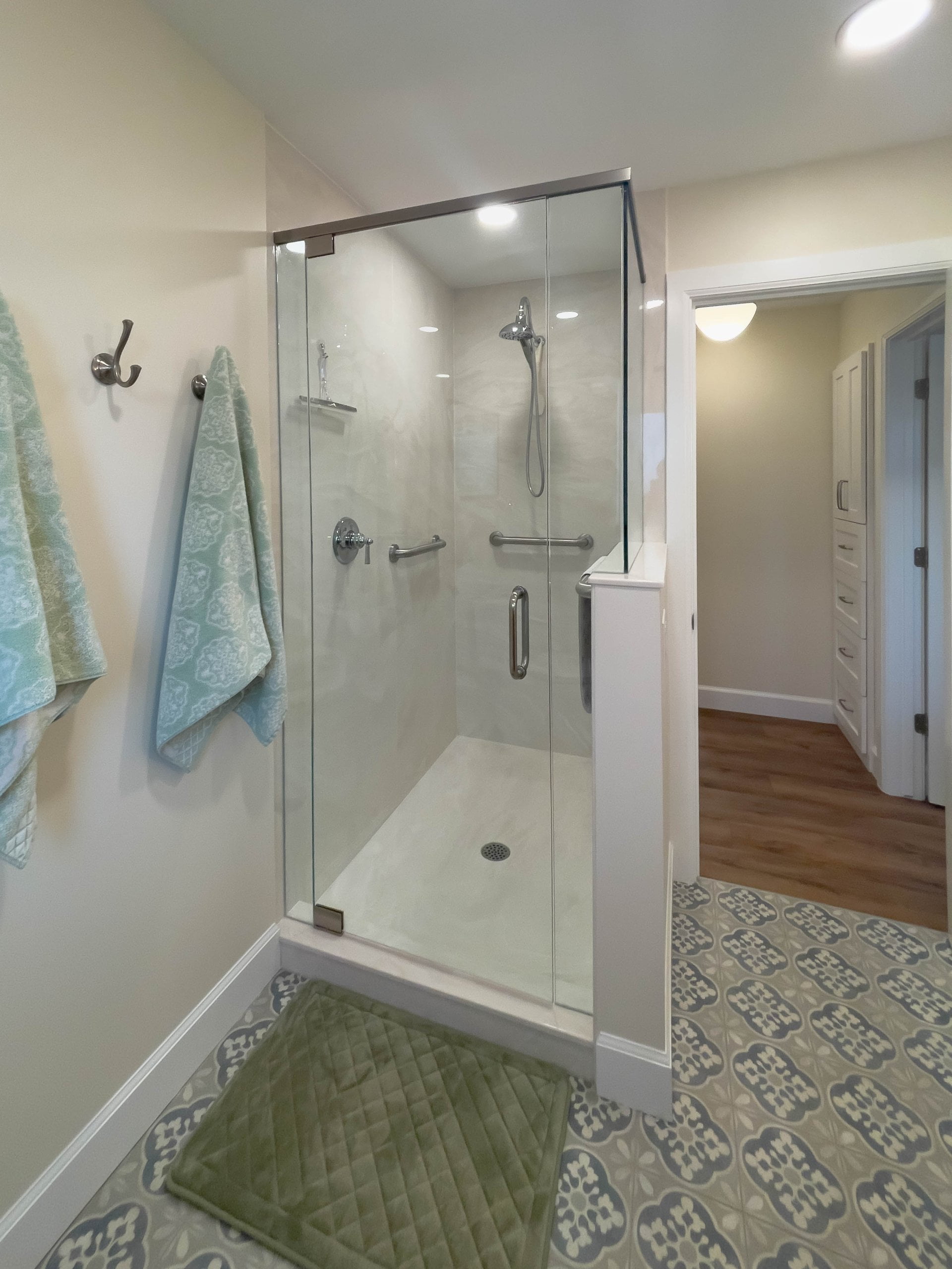 Shower with glass door. In front of the shower door is a green bath mat placed on top of the tile. Two towels are hung on the wall hooks to the side of the shower. The door to the bathroom is open and looks into the hallway.