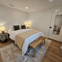 Thumbnail of http://Basement%20bedroom%20with%20a%20bed,%20bench,%20nightstand,%20and%20mirror%20hung%20on%20the%20door.