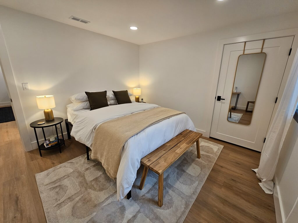 Basement bedroom with a bed, bench, nightstand, and mirror hung on the door.