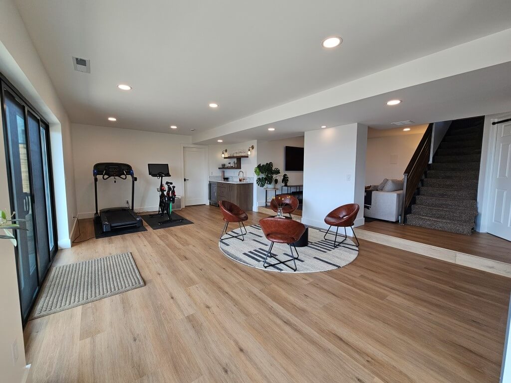 Large basement great room with four chairs, the stairs, a small bar, and a workout area.