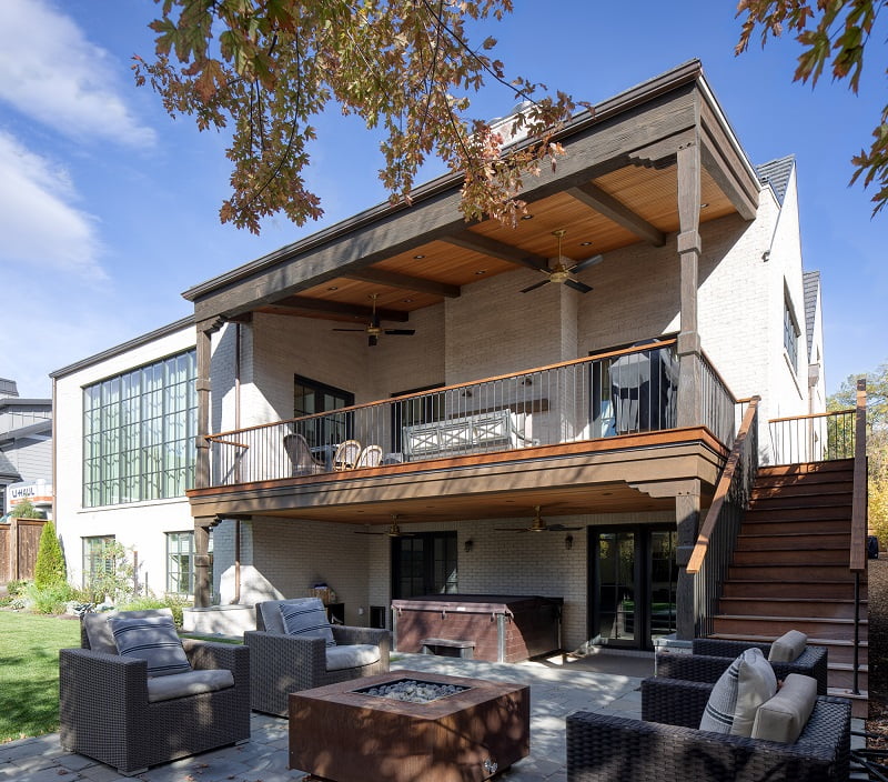 Backyard and outdoor entertaining space at a house. The lower level includes a partially covered patio with a hot tub and furniture around a fire pit. Upstairs, a covered deck includes ceiling fans, outdoor furniture, and a modern railing. Both levels have doors to the interior and are connected by a staircase on the right side.