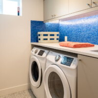 Thumbnail of http://Basement%20laundry%20room%20with%20a%20small%20window,%20blue%20tile%20backsplash,%20and%20side%20by%20side%20washer%20and%20dryer%20tucked%20under%20the%20counter.