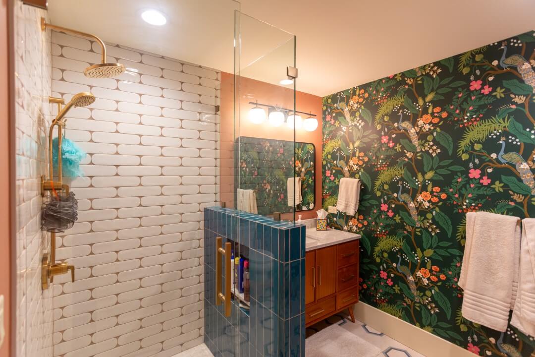 Renovated bathroom with white and blue tiles in the shower, gold fixtures, and a wallpaper that features peacocks.
