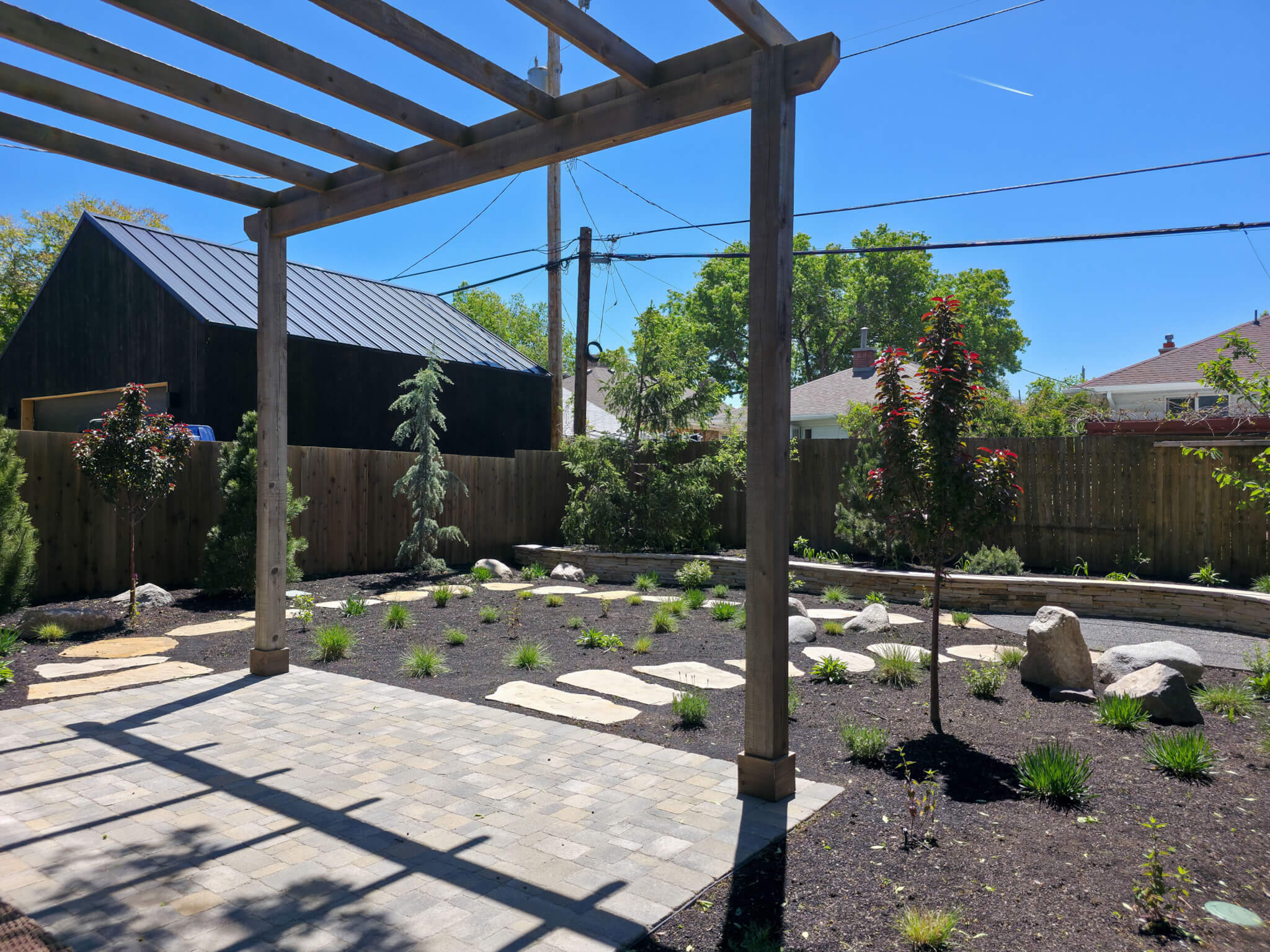 Freshly landscaped backyard. The yard includes a patio with a pergola, stone pathways, retaining wall, and various plants.