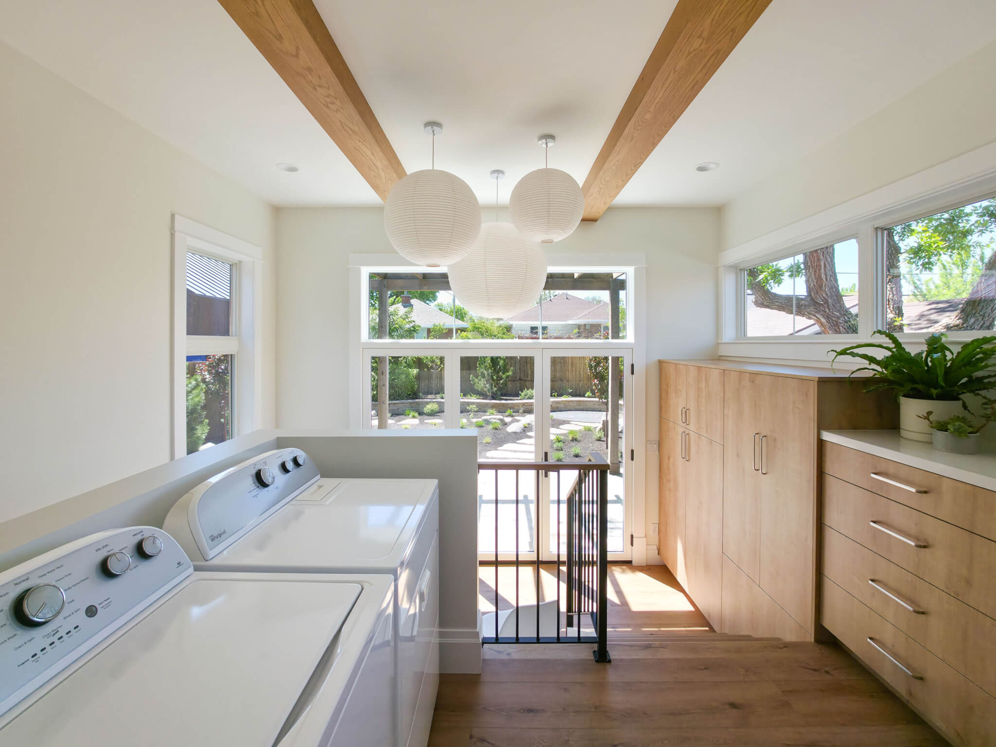 Laundry room section of the addition looking toward the back door in the mudroom section. Along the left is the washer and dryer, and along the right are built-in cabinets. The space features lots of natural light, wood beams on the ceiling, and three round light fixtures.