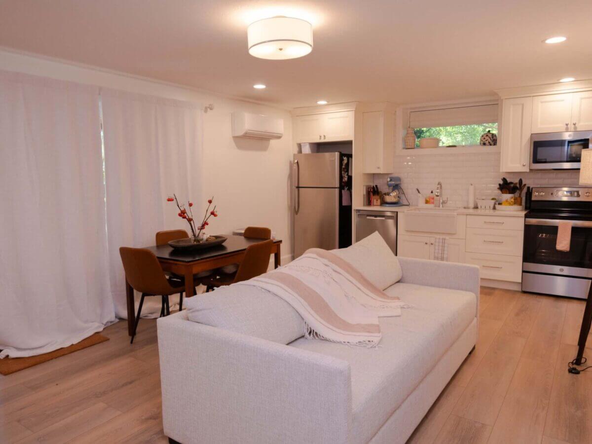 Basement apartment with a living room, dining space, and kitchen along the wall. The kitchen includes a fridge and freezer, dishwasher, farmhouse sink, and stove and oven with a microwave above.