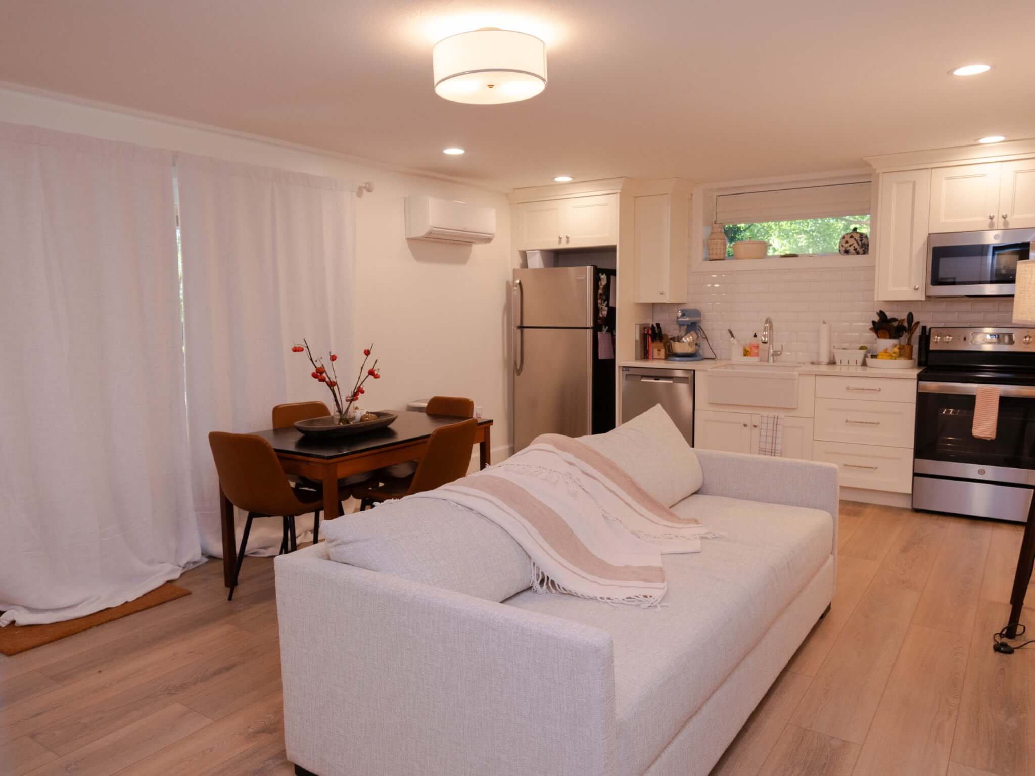 Basement apartment with a living room, dining space, and kitchen along the wall. The kitchen includes a fridge and freezer, dishwasher, farmhouse sink, and stove and oven with a microwave above.