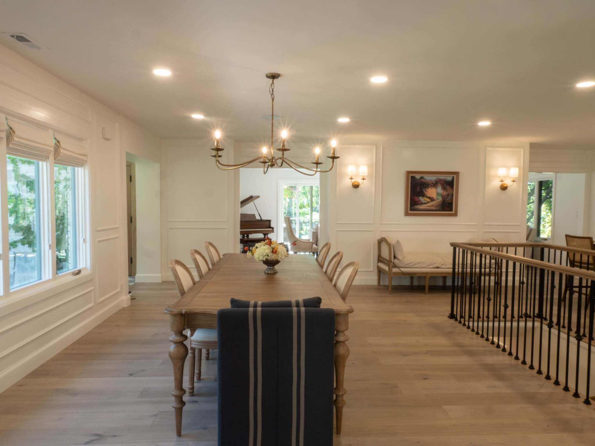 Open concept dining room with a table and eight chairs. Straight ahead is a view to the living room. On the left are windows. On the right is a modern railing around the staircase.