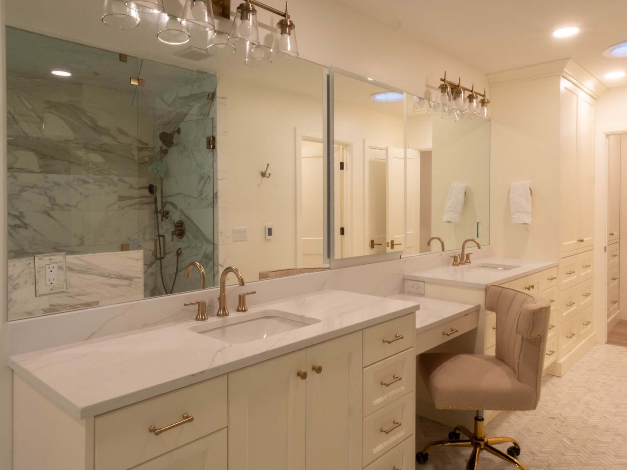 Primary bathroom vanity. The vanity includes two sinks with a makeup vanity between, Above the sinks are large mirrors and light fixtures.