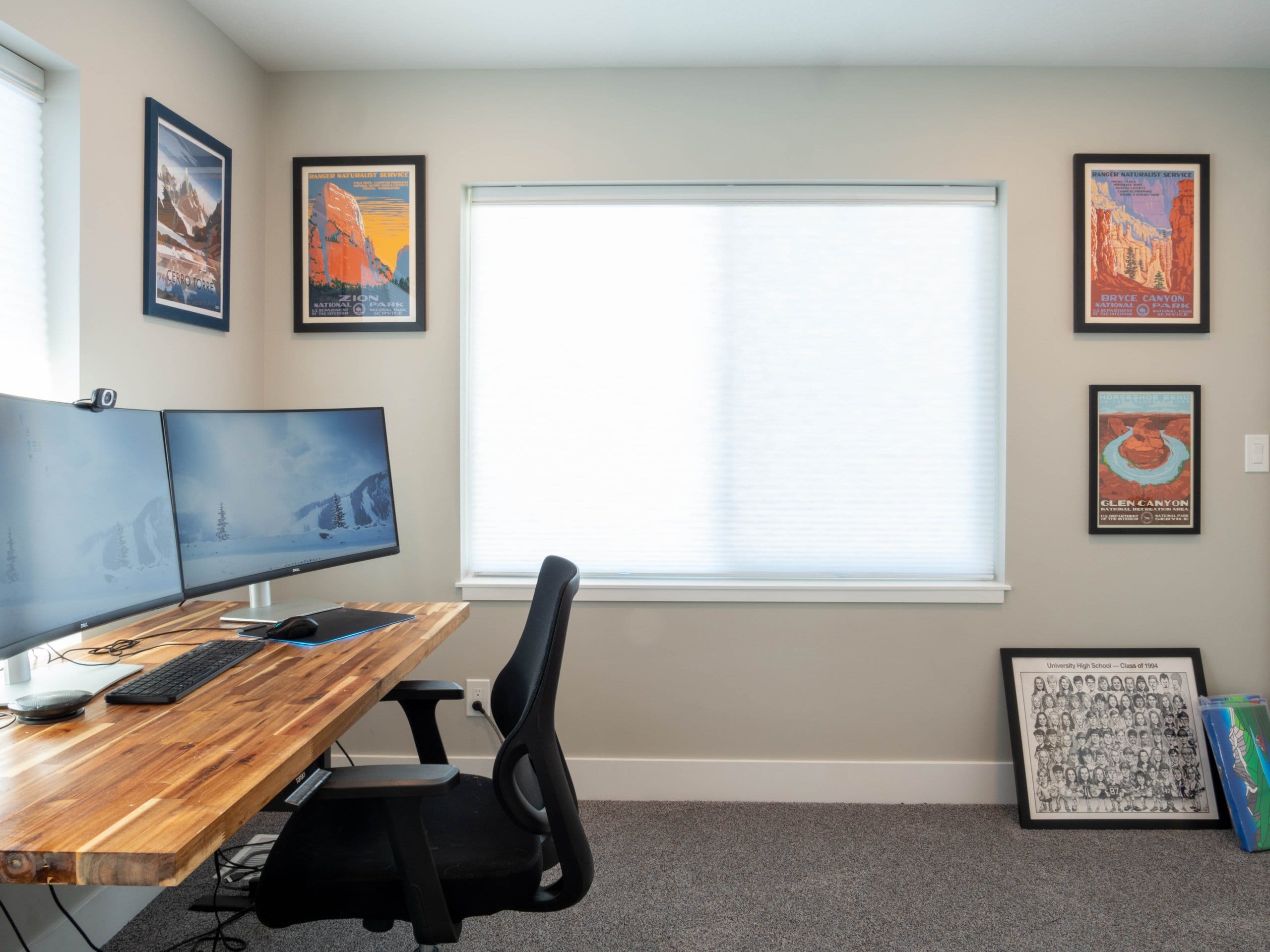 Carpeted office with a large window, pictures hung on the wall, and a desk that has two monitors.