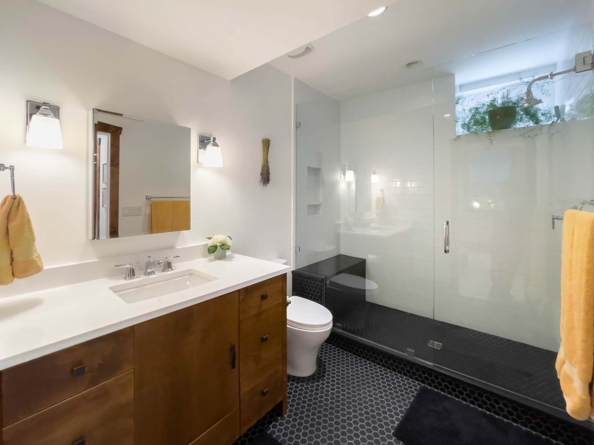 Bathroom with shower, toilet, and large sink. The shower includes a windowsill where a plant is as well as a niche and bench. The sink includes a mirror with lights hung on either side and storage below.