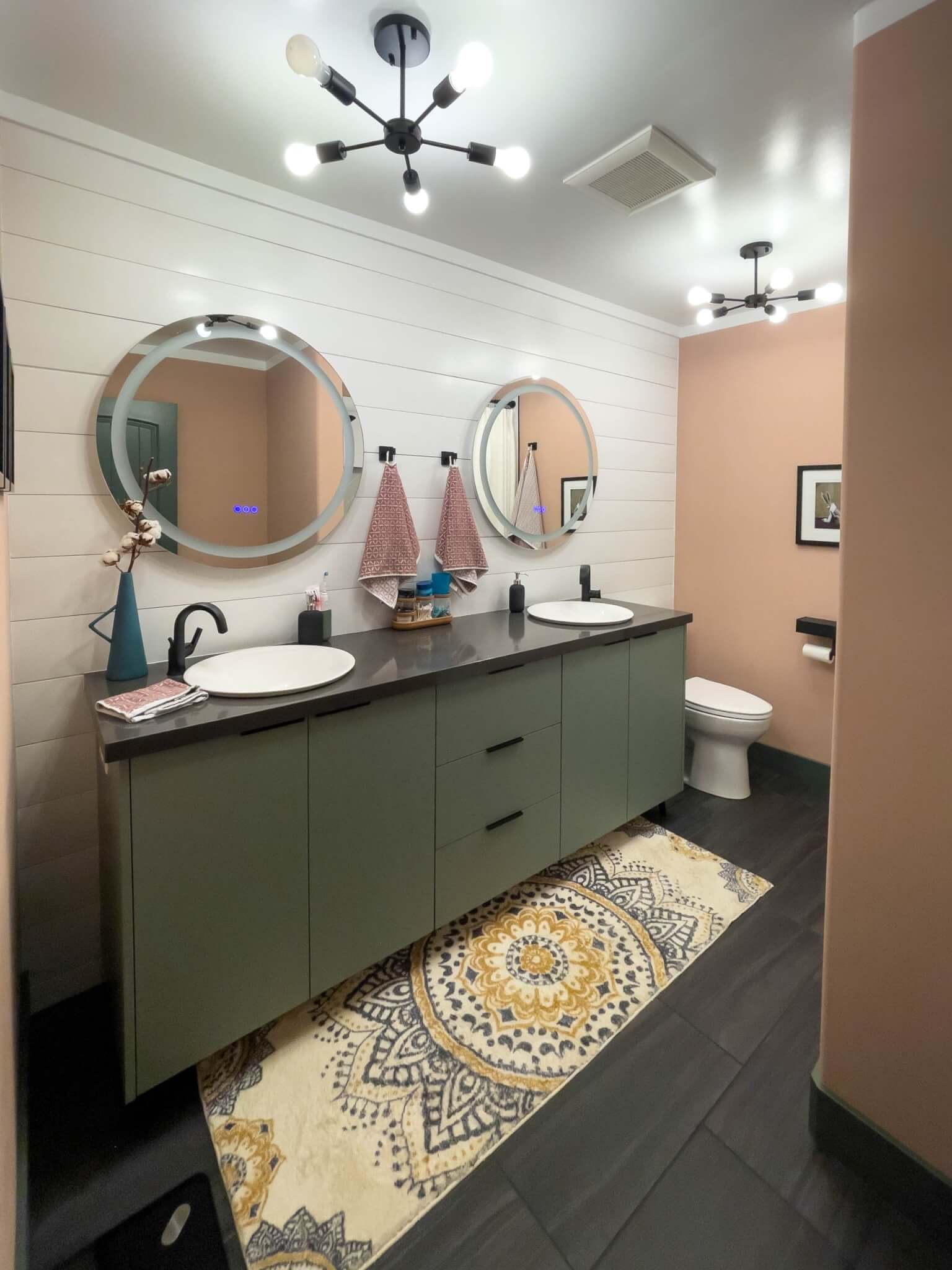 Primary bathroom looking at the double vanity and toilet. The vanity has a long rug in front, includes storage under each sink, and has mirrors with lights above. On the ceiling are two light fixtures and a fan.
