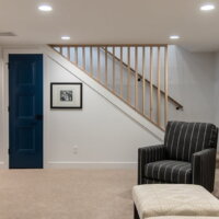 Thumbnail of http://Basement%20family%20room%20with%20a%20modern%20wood%20railing,%20small%20door%20to%20storage%20underneath,%20carpet,%20and%20furniture%20that%20includes%20an%20arm%20chair.