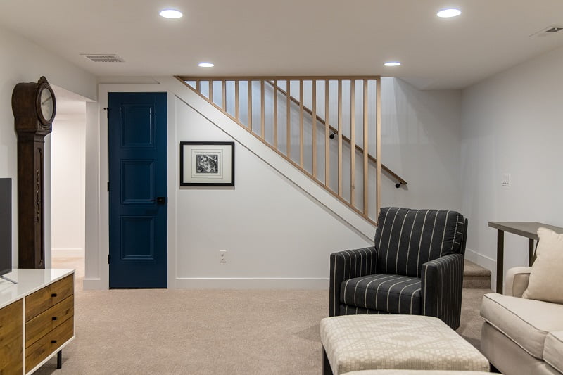 Basement family room with a modern wood railing, small door to storage underneath, carpet, and furniture that includes an arm chair.