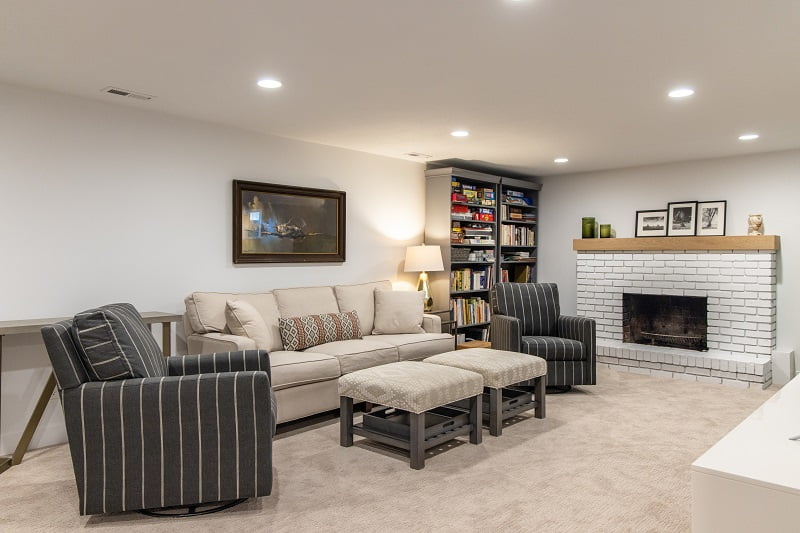 Contemporary basement remodel at a rambler home. The room features two arm chairs, two ottomans, a couch with decorative pillows, bookcases, and a white painted brick fireplace.