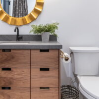 Thumbnail of http://Modern%20bathroom%20renovation.%20The%20toilet%20is%20on%20the%20right%20and%20the%20wood%20vanity%20is%20on%20the%20left.%20Between%20a%20roll%20of%20toilet%20paper%20is%20mounted%20to%20the%20vanity%20and%20a%20waste%20basket%20sits%20on%20the%20tile%20floor.%20Above%20the%20sink%20is%20a%20round,%20gold%20mirror.