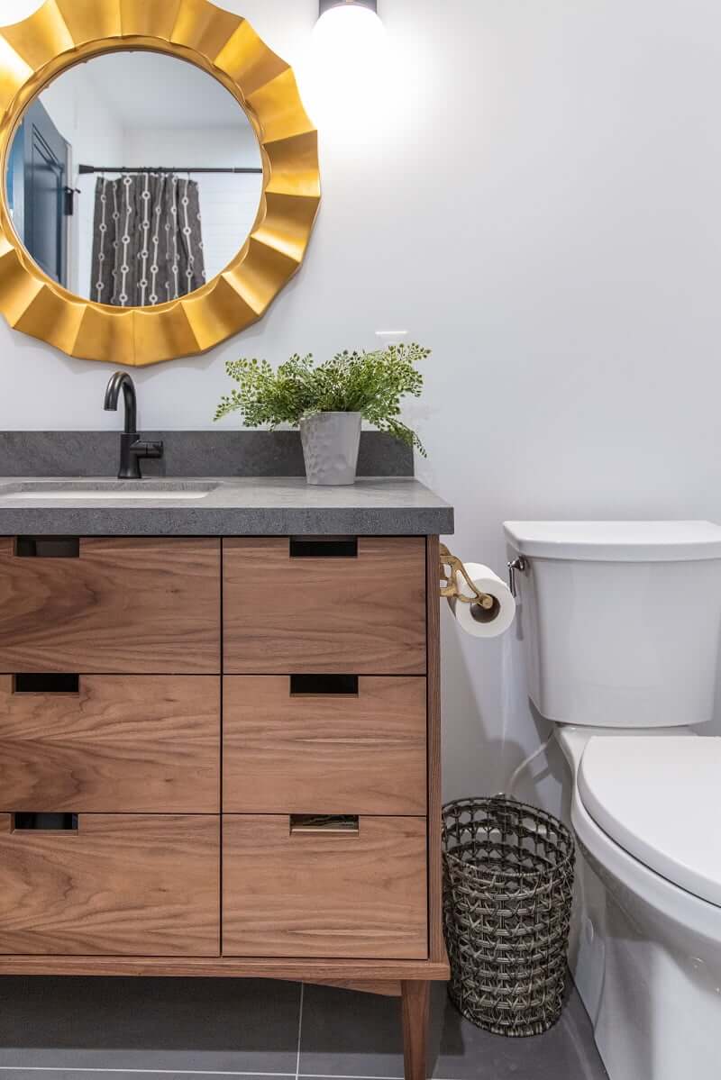 Modern bathroom renovation. The toilet is on the right and the wood vanity is on the left. Between a roll of toilet paper is mounted to the vanity and a waste basket sits on the tile floor. Above the sink is a round, gold mirror.